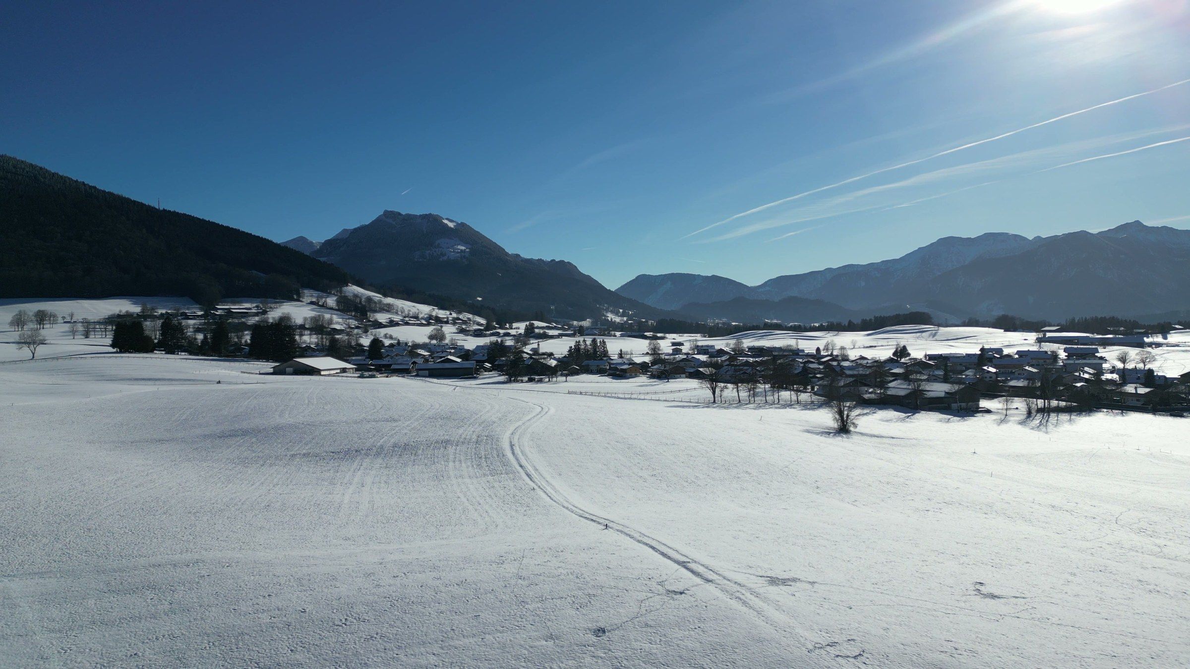 Drohnenblick über verschneite Wiesen mit einer Loipenspur auf Hundham, dahinter ein bewaldeter Berg und Bergketten unter blauem Himmel