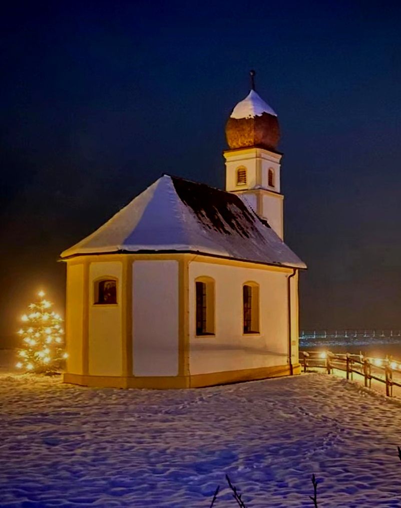 Die angestrahlte Leonhardi-Kapelle mit Zwiebelturm und Schnee auf Dach und Turm unter dunkelblauem Abendhimmel, links ein Christbaum mit Lichtern, rechts hinten ein Holzzaun mit Lichterkette, vorne verschneite Wiese