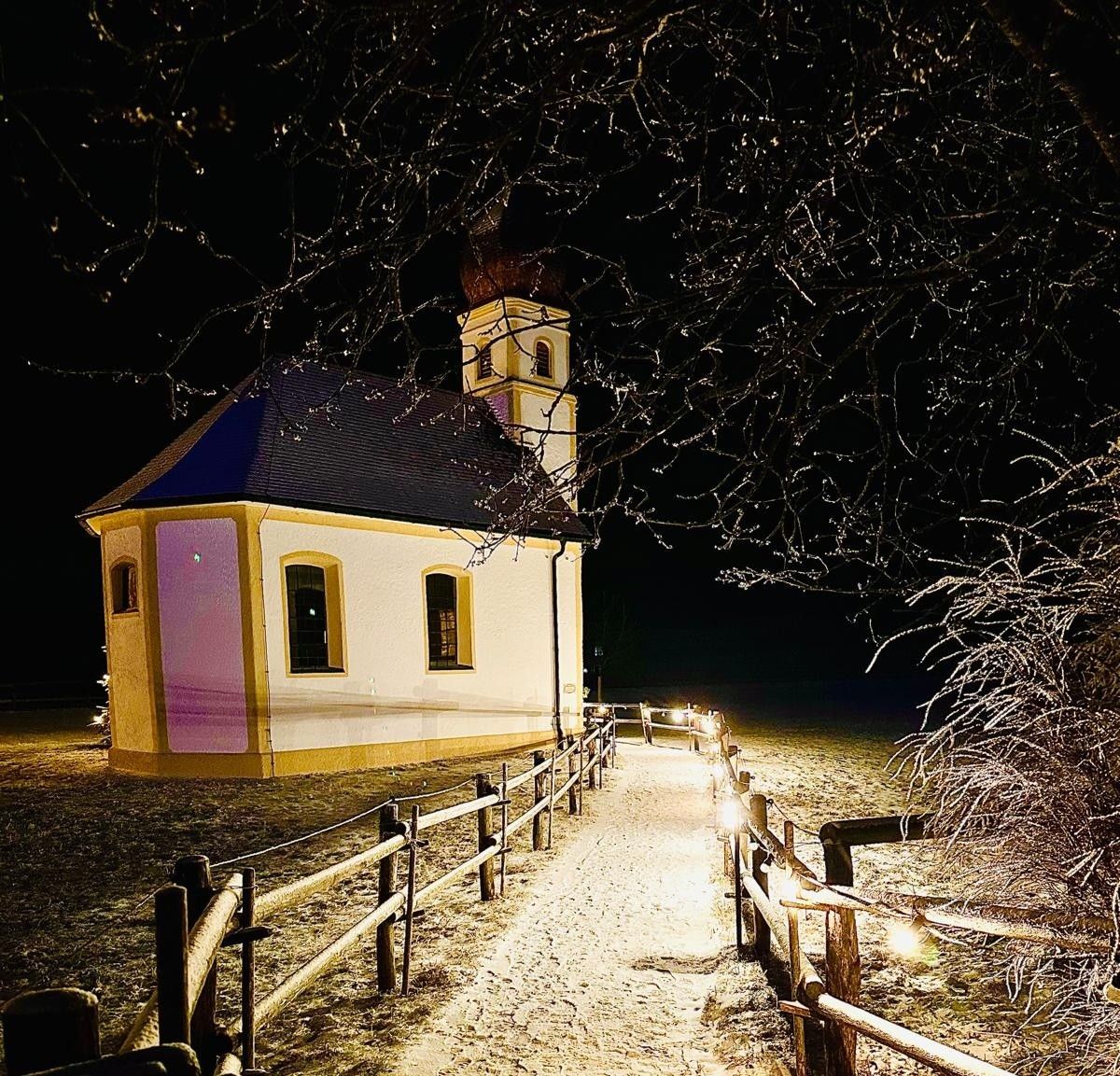 Ein verschneiter Weg zwischen zwei Holzzäunen mit Lichterketten führt nachts zur angestrahlten Leonhardi-Kapelle, oben und rechts die Äste eines Baums mit Raureif vor schwarzem Himmel