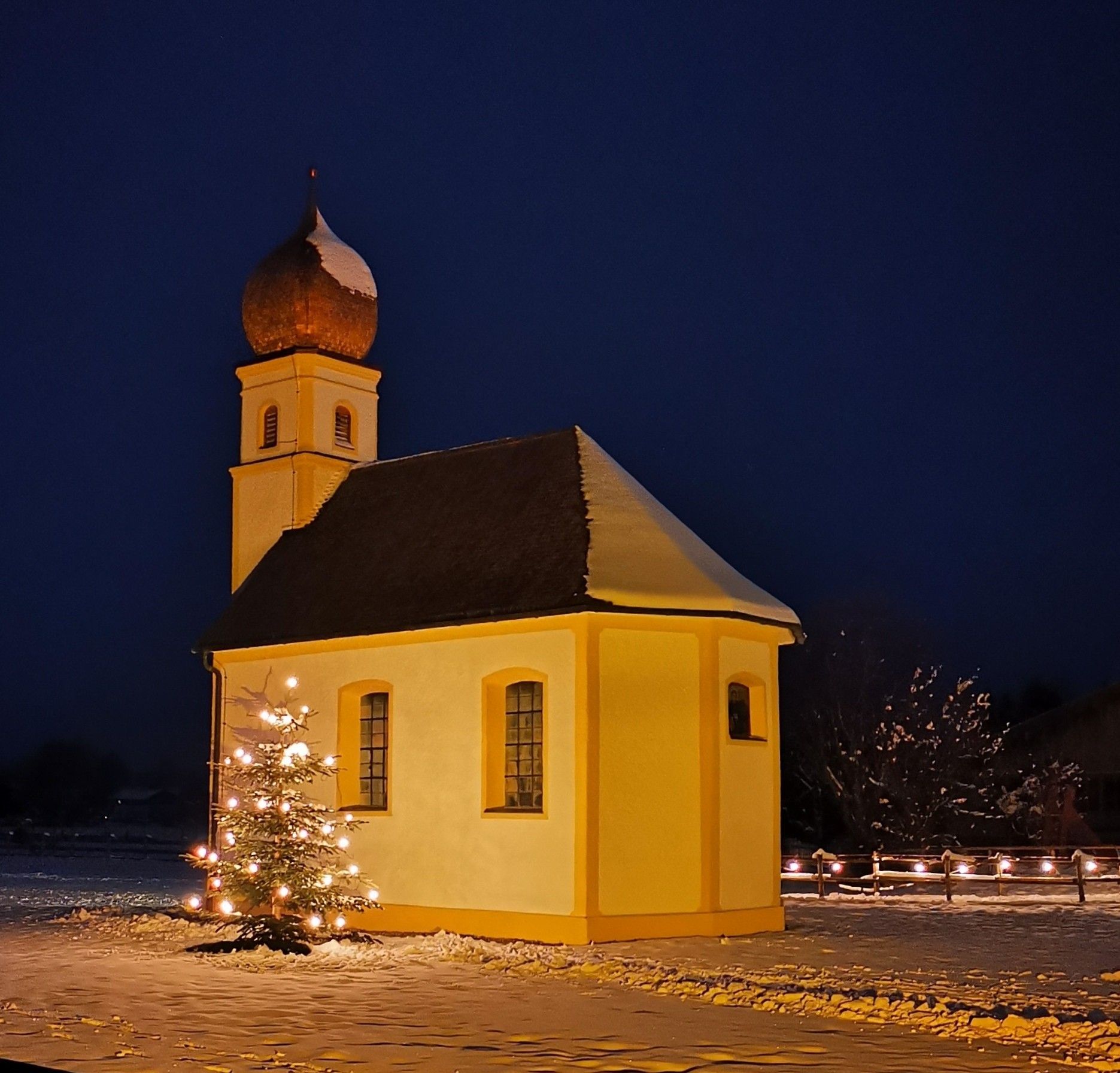 Die angestrahlte Leonhardi-Kapelle aus der Nähe an einem Winterabend, mit Zwiebelturm und Schnee auf dem Dach, links daneben ein Christbaum mit Lichtern, rechts dahinter ein Holzzaun mit Lichterkette auf der verschneiten Wiese