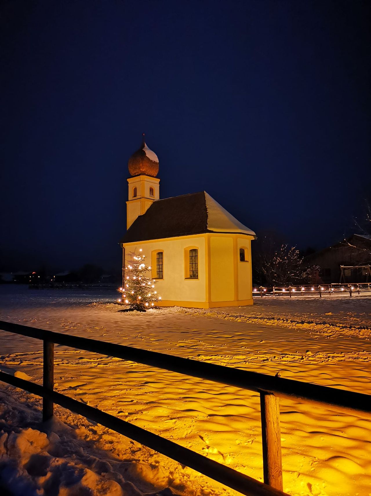 Die angestrahlte Leonhardi-Kapelle mit gelben Kanten und Zwiebelturm an einem Winterabend unter dunkelblauem Himmel, daneben ein Christbaum mit Lichtern, im Vordergrund ein Holzzaun vor goldgelb beleuchtetem Schnee