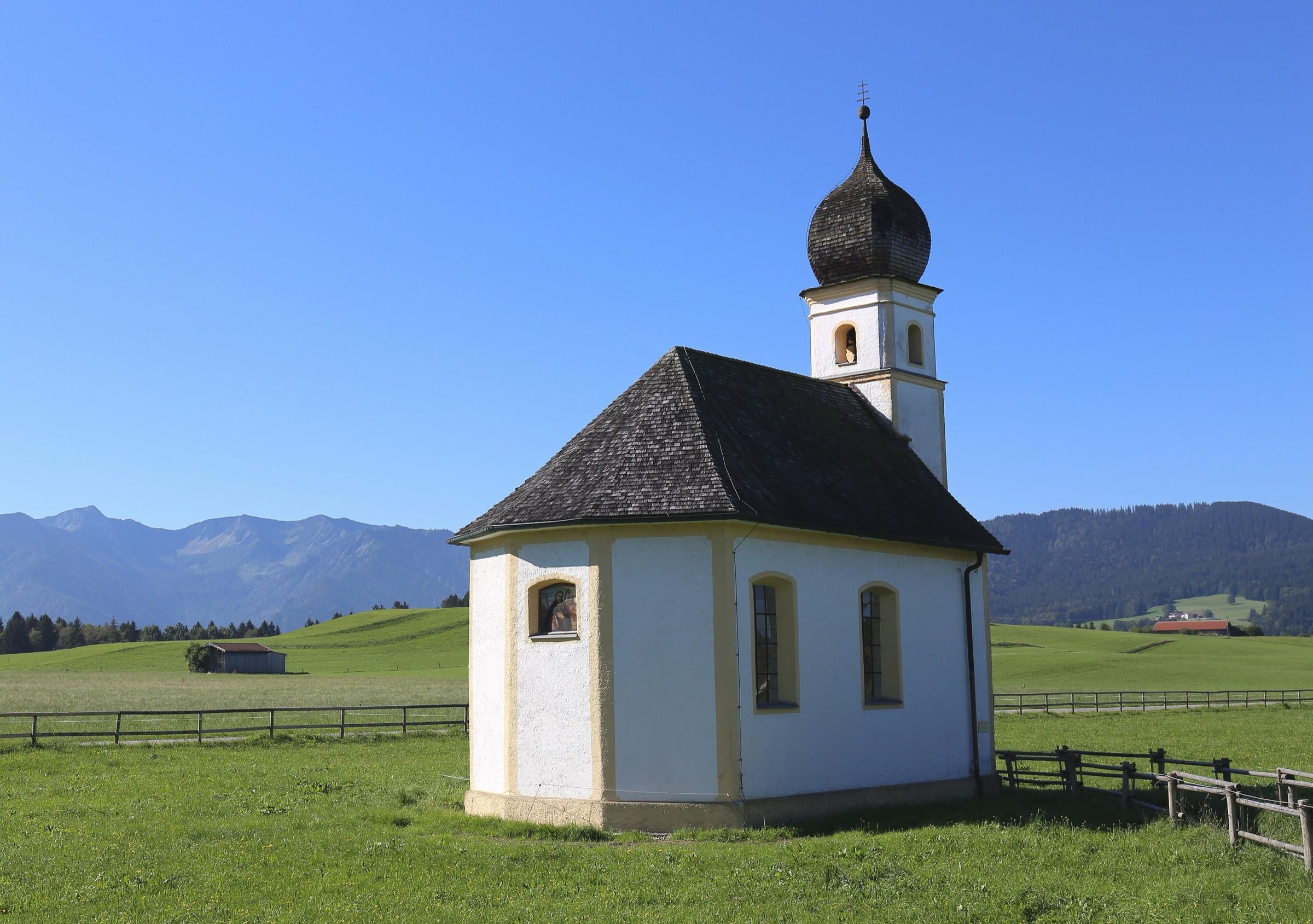 Weiße Kapelle mit Zwiebelturm auf einer eingezäunten Weide, dahinter die Voralpen unter blauem Himmel