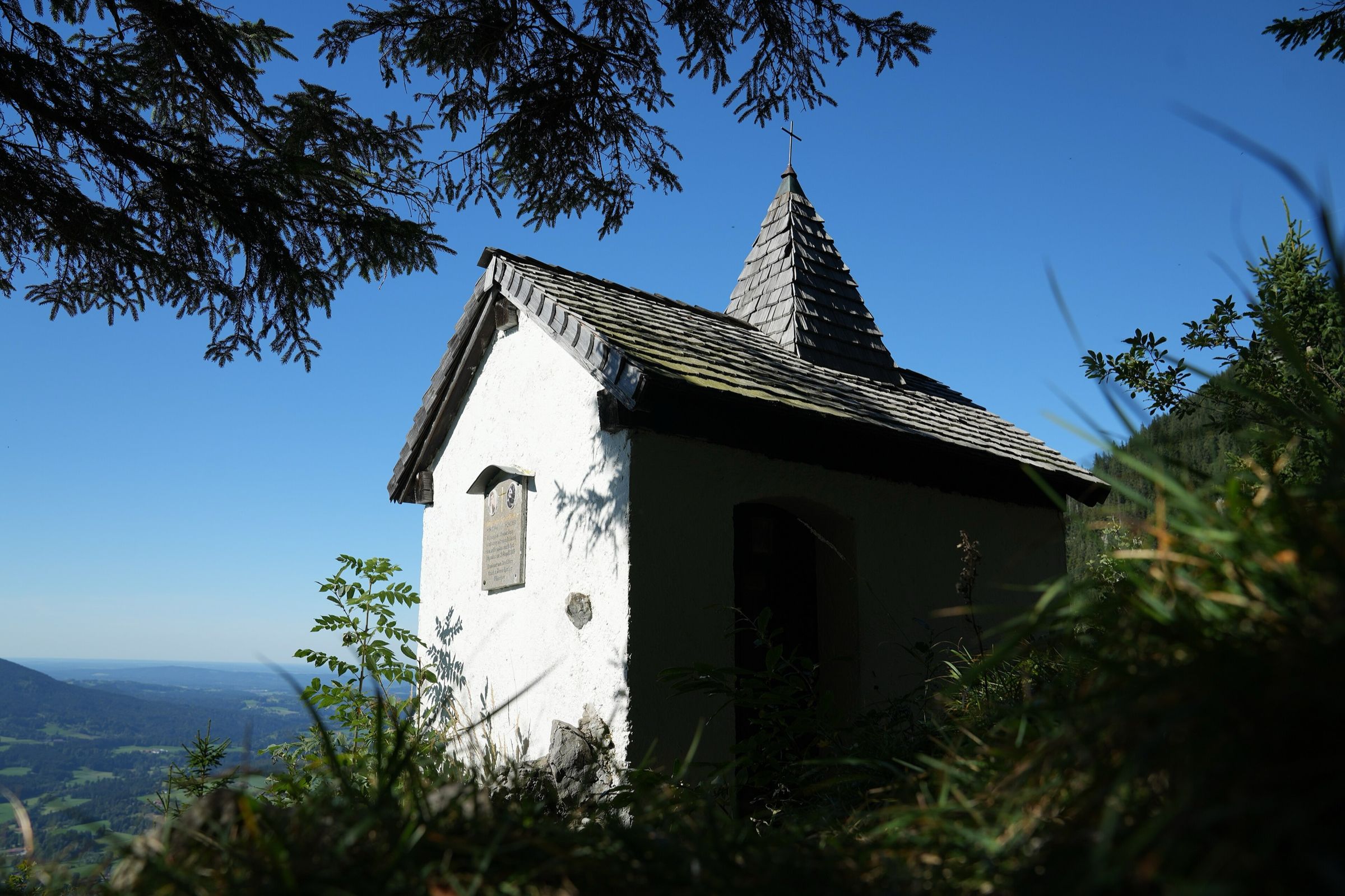 Kleine weiße Kapelle mit Schindeldach im Gegenlicht, umrahmt von Zweigen, im Hintergrund das Tal