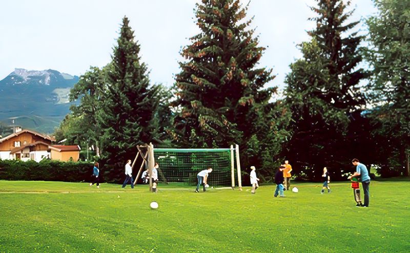 Kinder und Erwachsene spielen Fußball auf einer großen Rasenfläche mit Tor vor hohen Fichten, links ein Haus und ein Berg