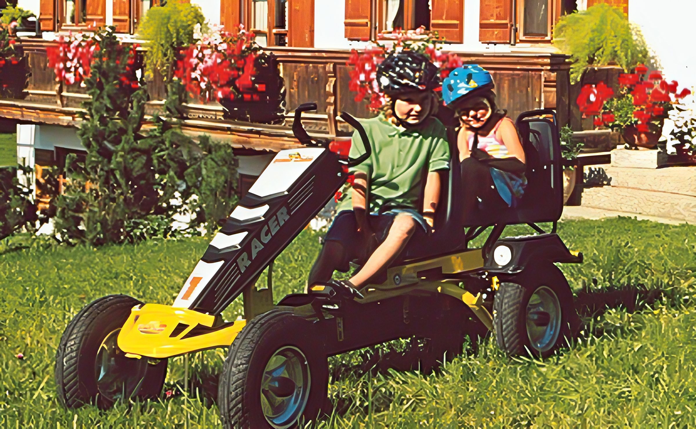 Zwei Kinder mit Fahrradhelm auf einem Kettcar im Gras, dahinter ein Holzbalkon mit roten Geranien