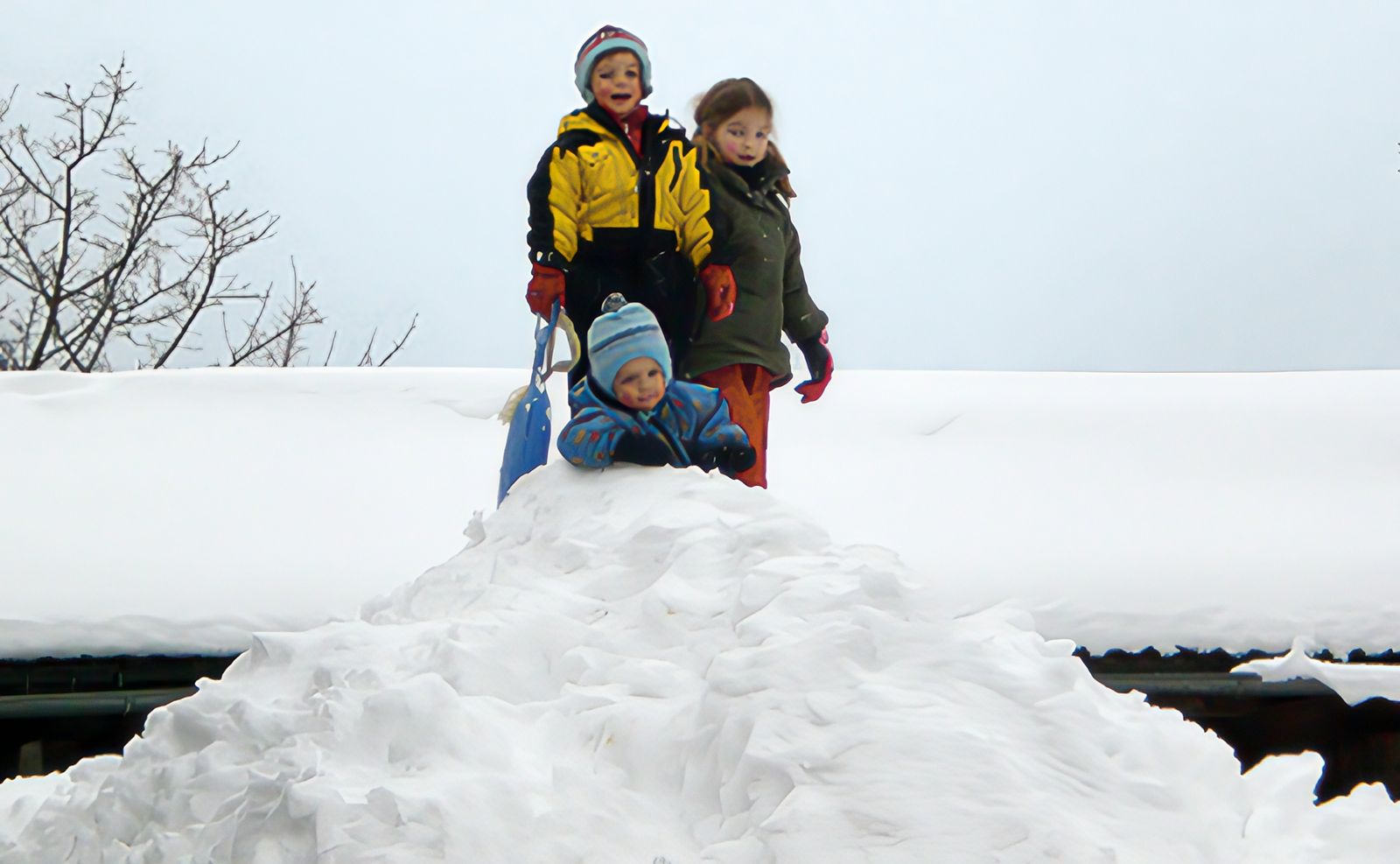 Drei Kinder in Winterkleidung stehen und sitzen oben auf einem großen Schneehaufen vor einem verschneiten Dach