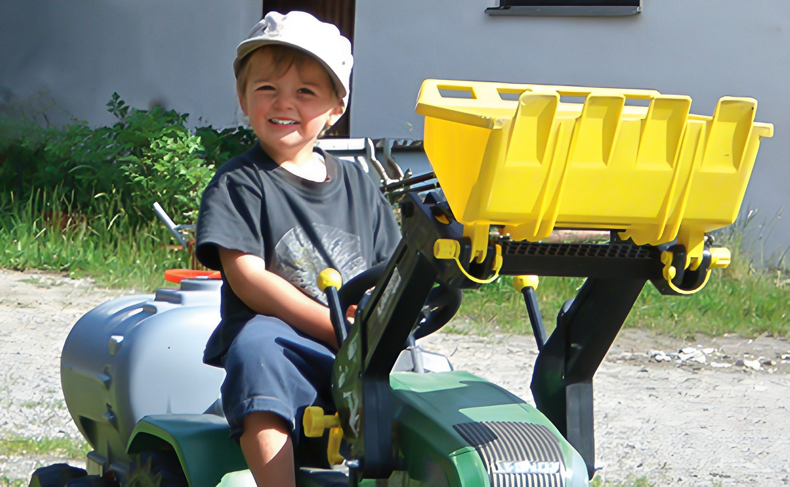 Lachendes Kleinkind mit weißer Mütze auf einem grünen Spieltraktor mit gelber Frontladerschaufel