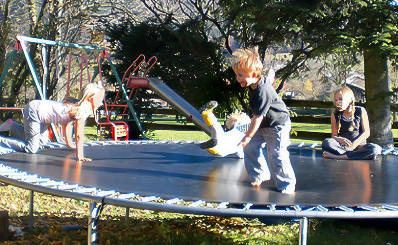 Vier Kinder spielen in der Herbstsonne auf einem großen Trampolin, dahinter Schaukel, Rutsche und Bäume