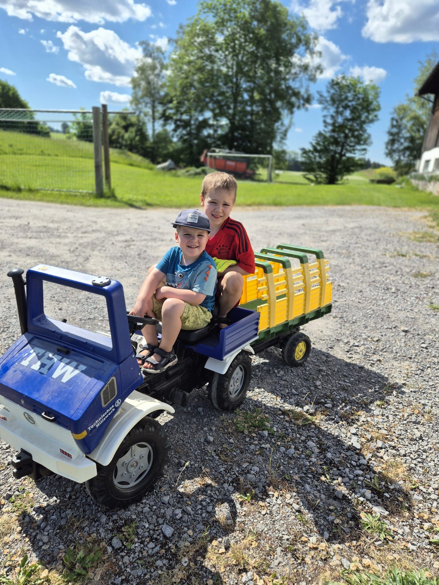 Two children sitting on a blue pedal Unimog with a yellow trailer on the gravel yard in front of the farm, the meadow with the football goal and trees behind under a blue sky