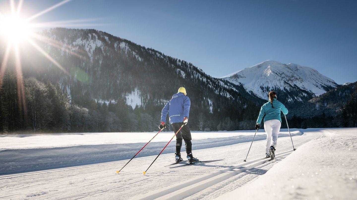 Zwei Langläufer von hinten in der gespurten Loipe auf einer verschneiten Talwiese, links ein bewaldeter Berg im Gegenlicht der Sonne, rechts ein schneebedeckter Gipfel unter blauem Himmel