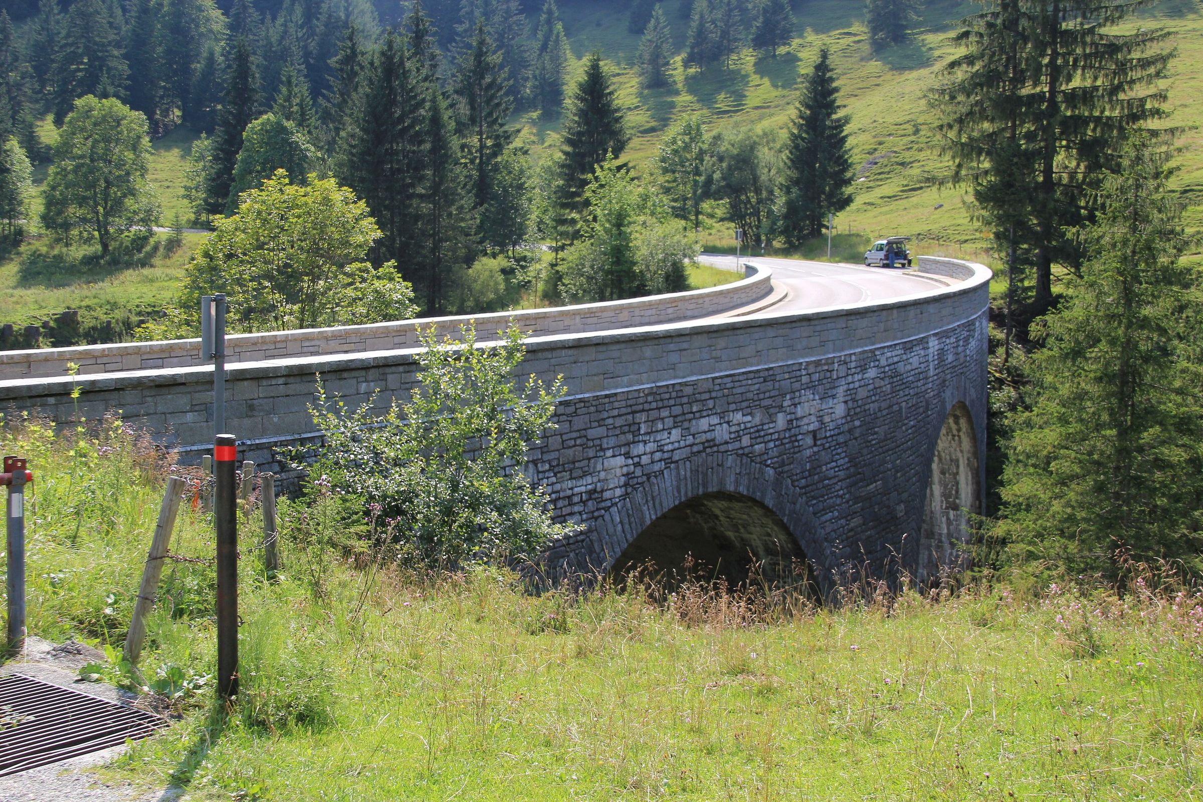 Geschwungene Steinbogenbrücke der Passstraße zwischen Almwiesen und Fichten, vorne ein Wegweiserpfosten, hinten ein Auto
