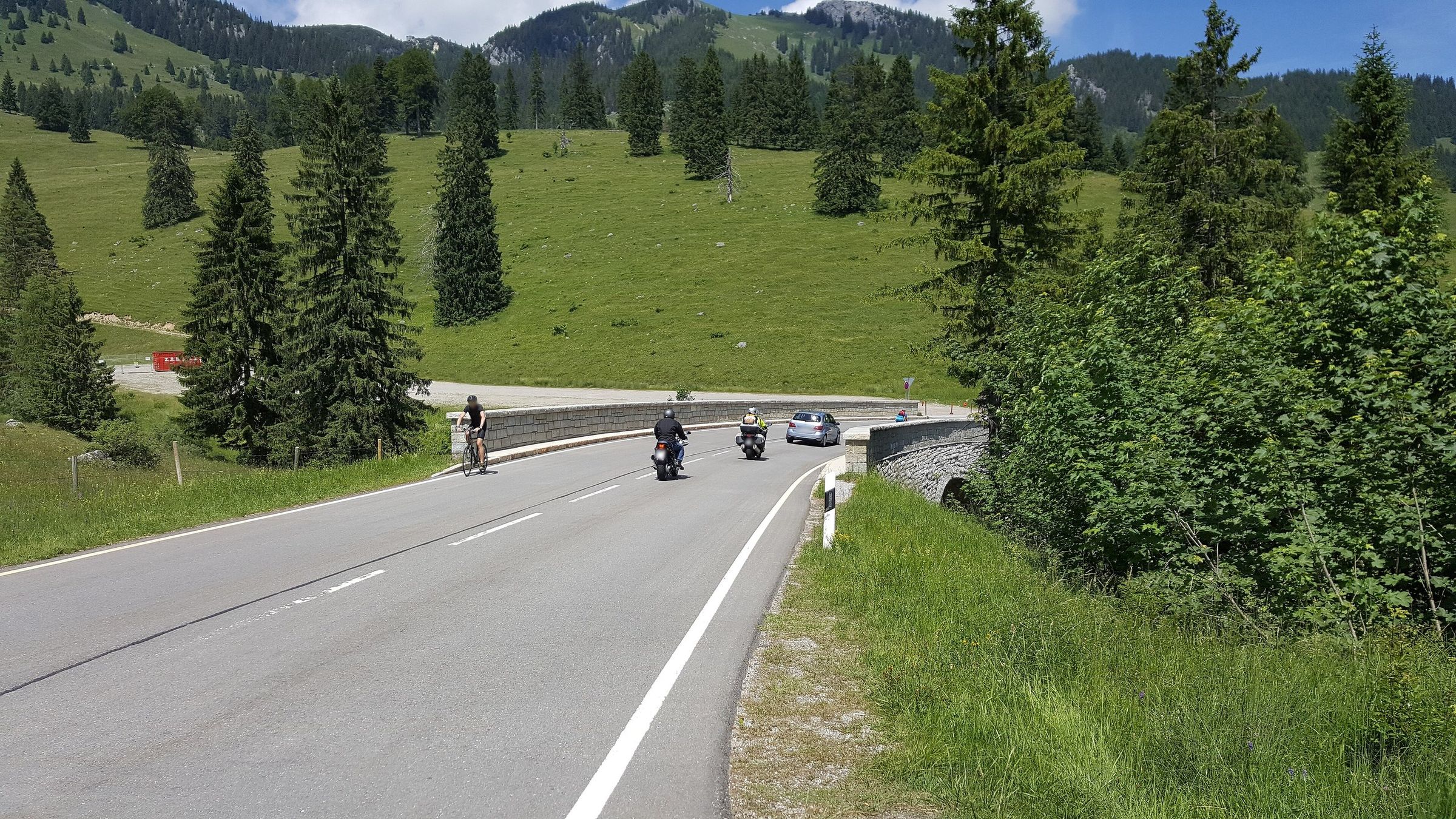Geschwungene Passstraße mit Steinmauer, darauf zwei Motorradfahrer von hinten, ein Rennradfahrer und ein Auto, dahinter Almwiesen mit Fichten und Berge