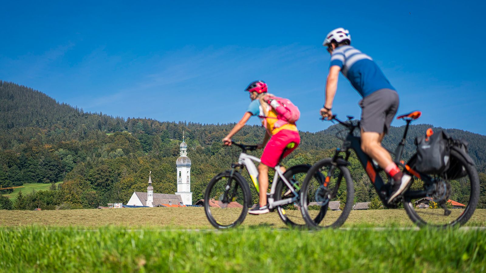 Zwei Radfahrer mit Helm auf E-Bikes fahren auf einem Wiesenweg, dahinter eine Kirche mit Zwiebelturm, Wald und blauer Himmel