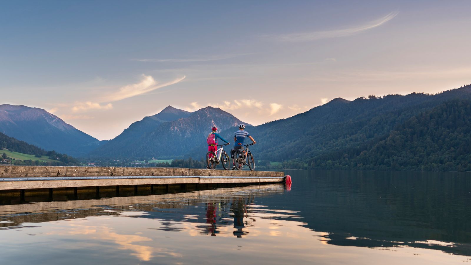 Zwei Radfahrer mit Helm stehen mit ihren Rädern am Ende eines Stegs auf einem stillen See, dahinter bewaldete Berge im Abendlicht