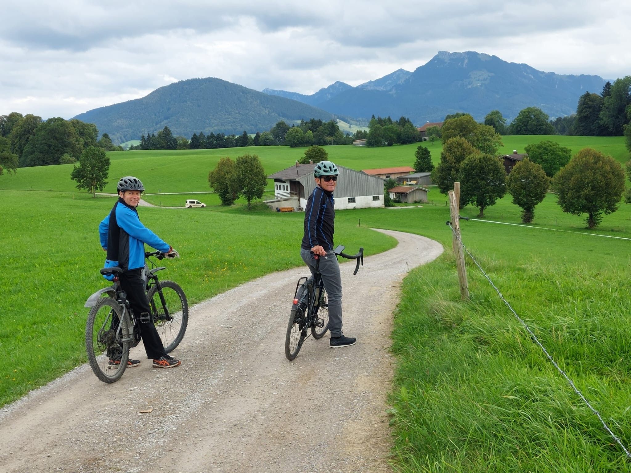Zwei Radfahrer mit Helm auf einem Schotterweg durch grüne Wiesen, dahinter ein Hof, Bäume und Berge