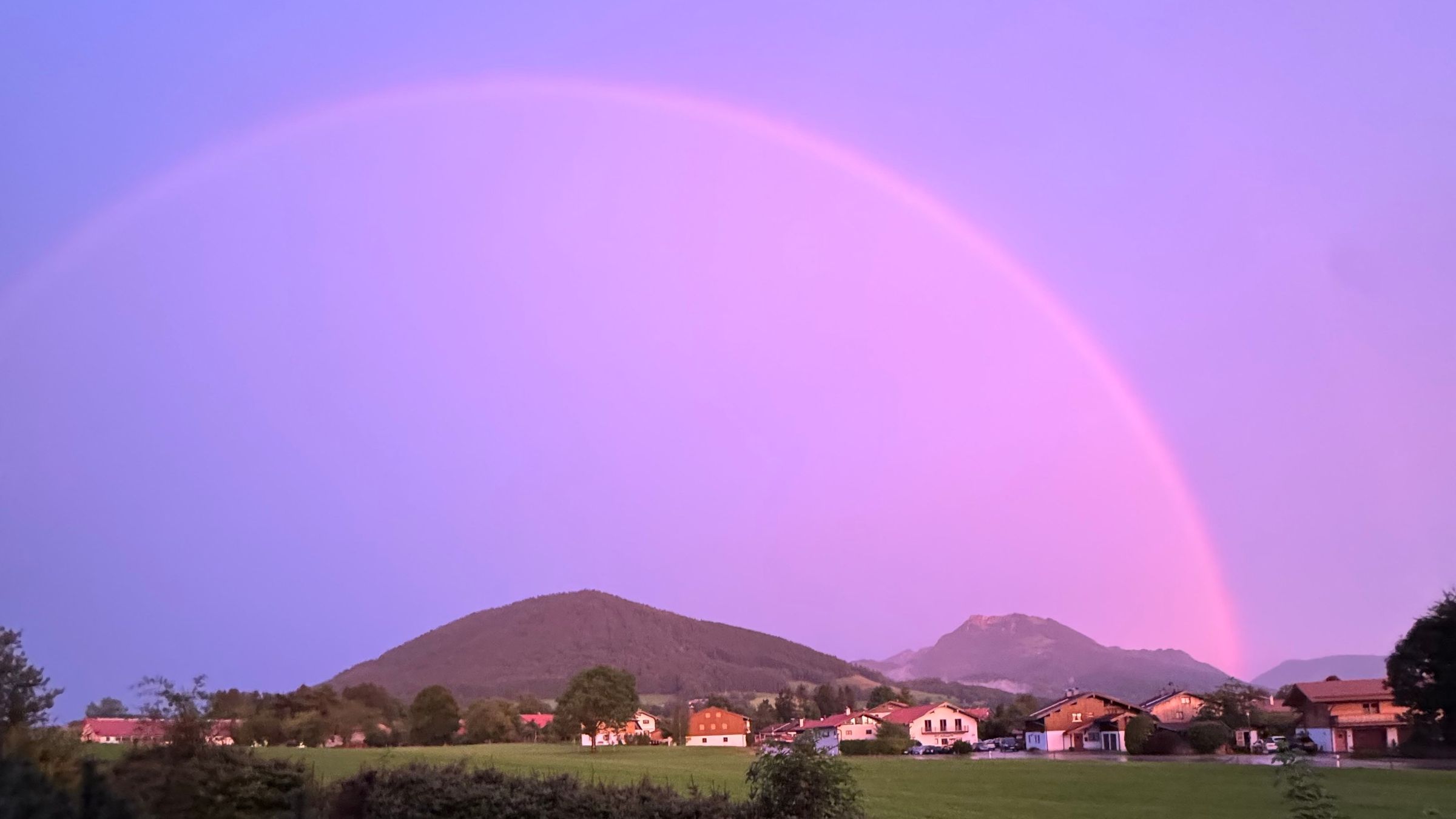 Regenbogen im violetten Abendlicht über Hundham, darunter Häuser, Wiesen und zwei Berge