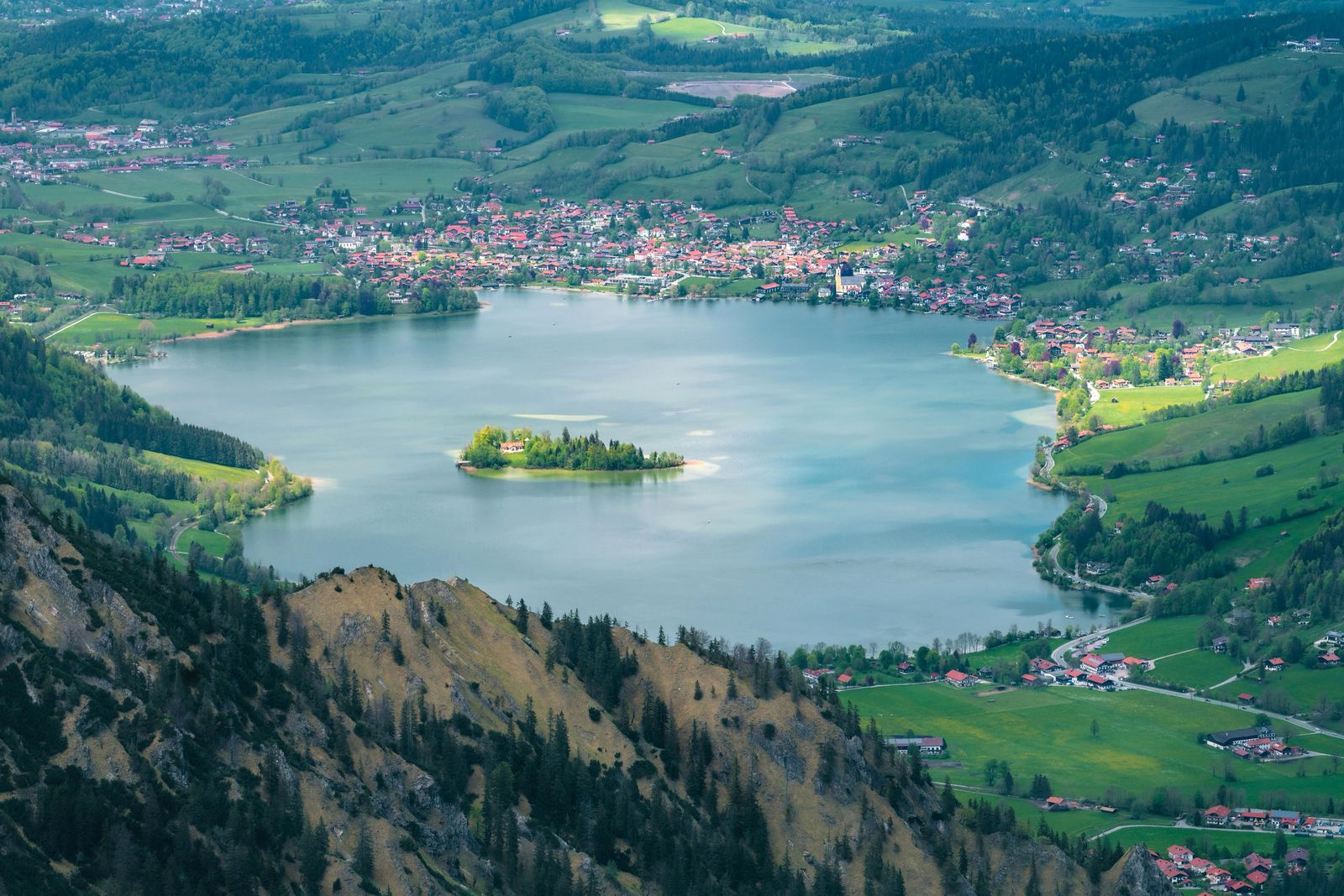 Blick von einem Berghang auf den ganzen Schliersee mit der Insel Wörth, dahinter der Ort Schliersee, Wiesen und das Alpenvorland