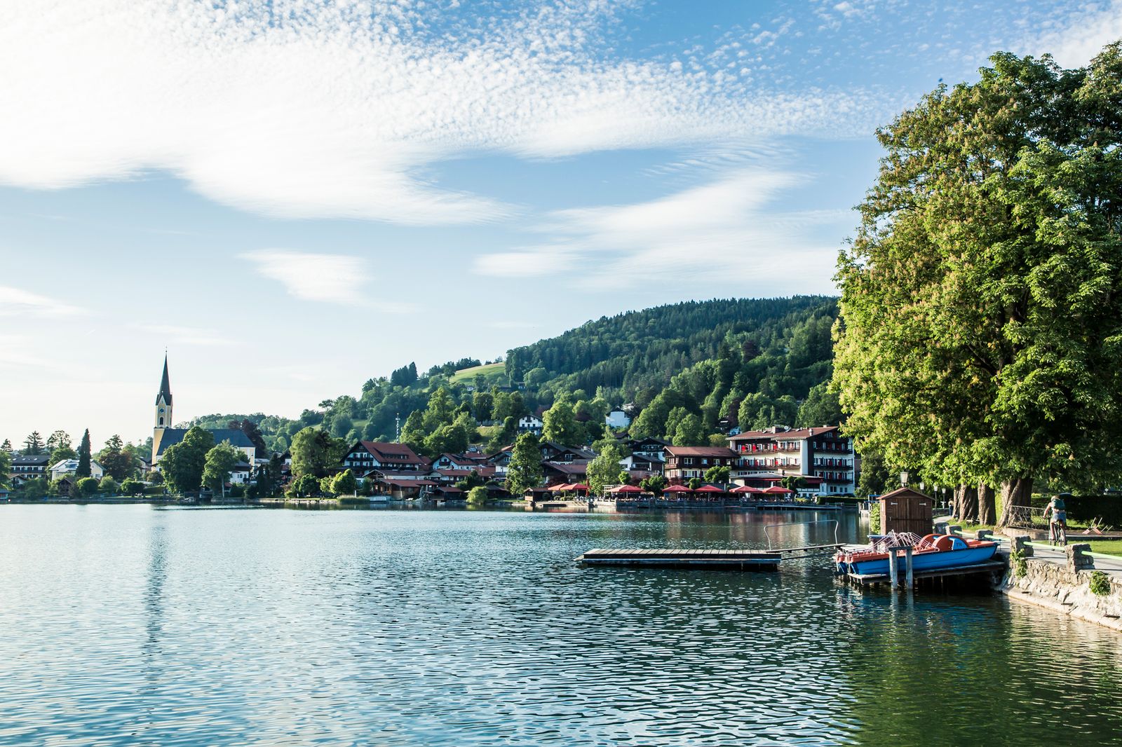 Der Ort Schliersee mit Kirchturm am Seeufer, davor glitzerndes Wasser, ein Steg und Boote, rechts große Bäume