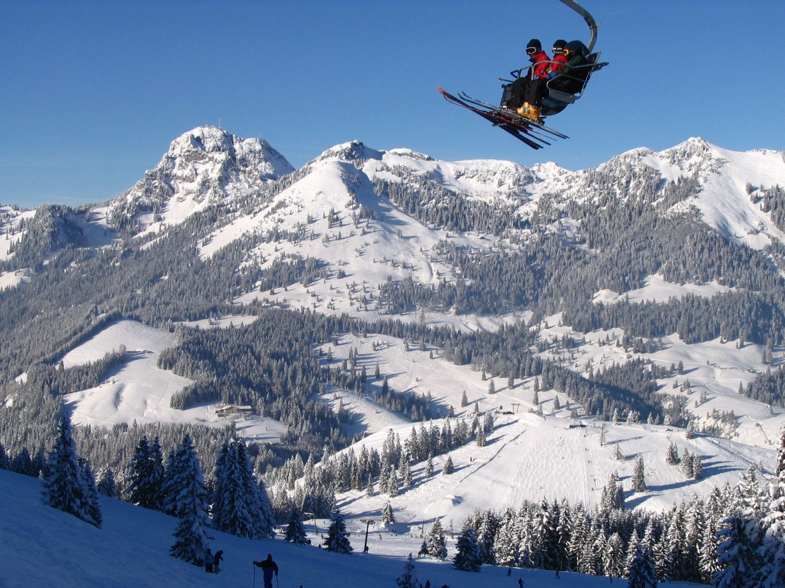 Drei Skifahrer mit Mützen und Skibrillen im Sessellift vor tiefblauem Himmel, darunter verschneite Pisten und Wälder des Sudelfelds, links der Wendelstein