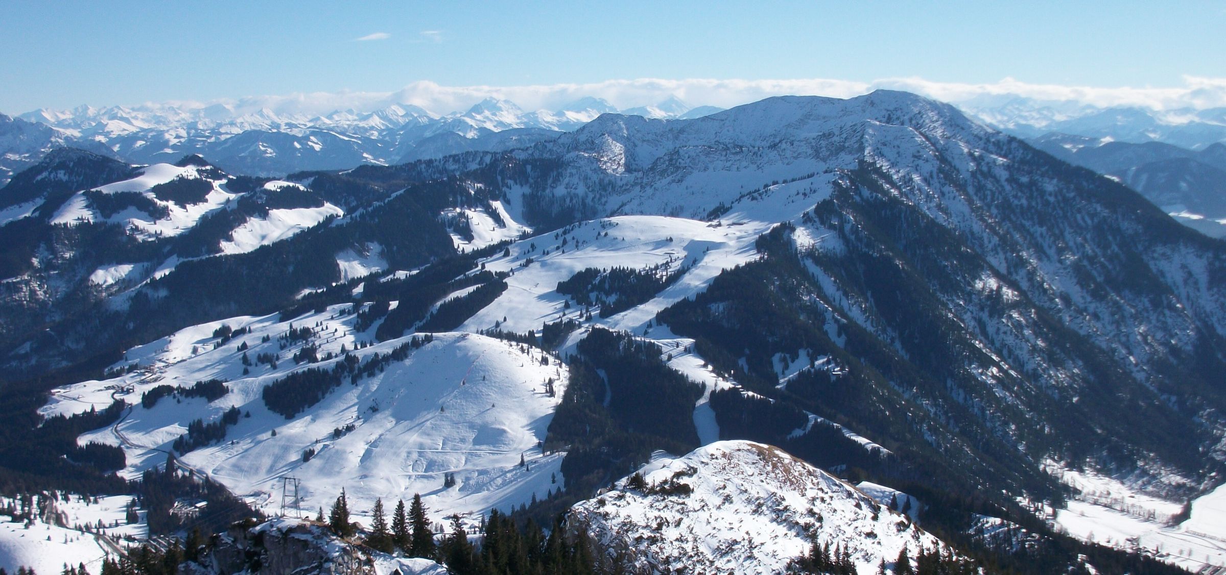 Verschneite Bergrücken des Sudelfelds mit Waldinseln, am Horizont die Alpen