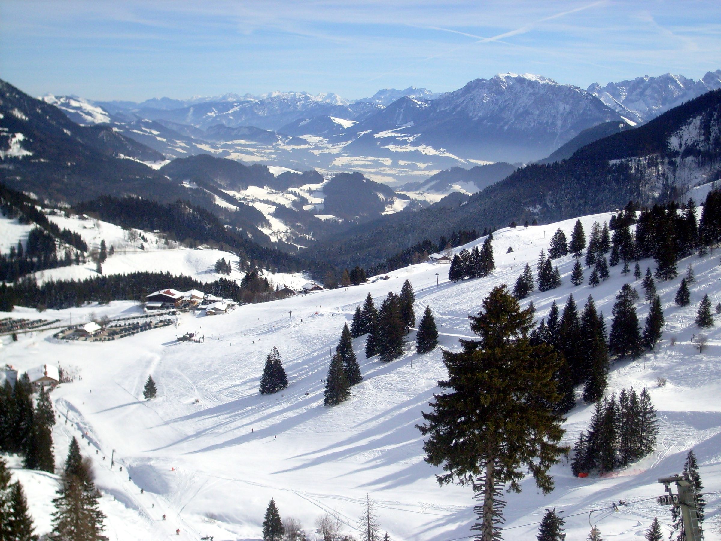 Sonnige Skipisten zwischen Fichten, unten Talstation und Parkplatz, dahinter das Inntal und Berge
