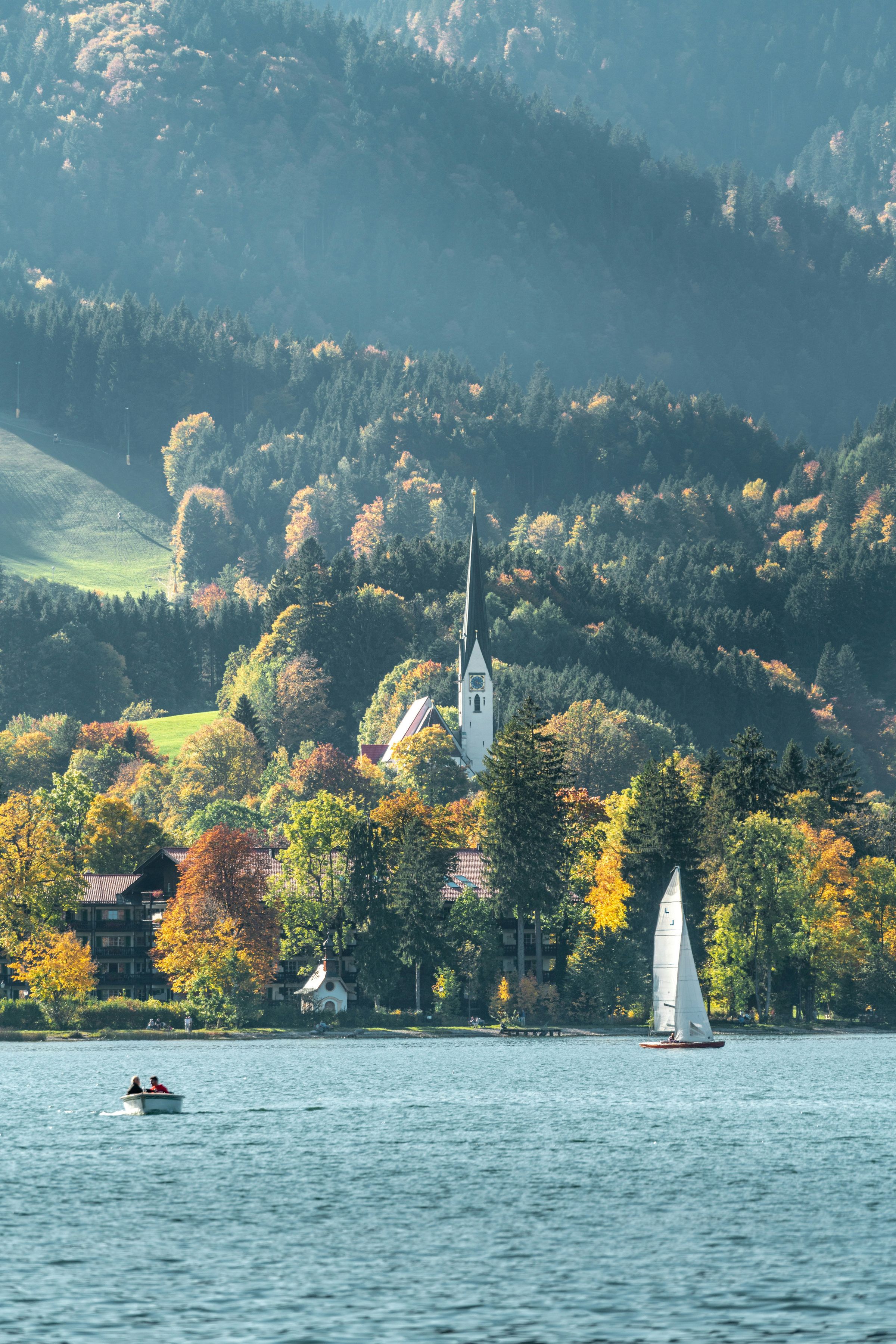 Weiße Kirche mit spitzem Turm am bewaldeten Ufer des Tegernsees, davor gelbe und rote Herbstbäume, Uferhäuser und ein weißes Segelboot auf dem See