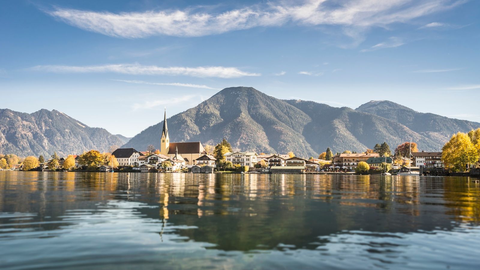 Kirche und Uferhäuser von Rottach-Egern spiegeln sich im Tegernsee, dahinter ein Berg, Herbstlaub