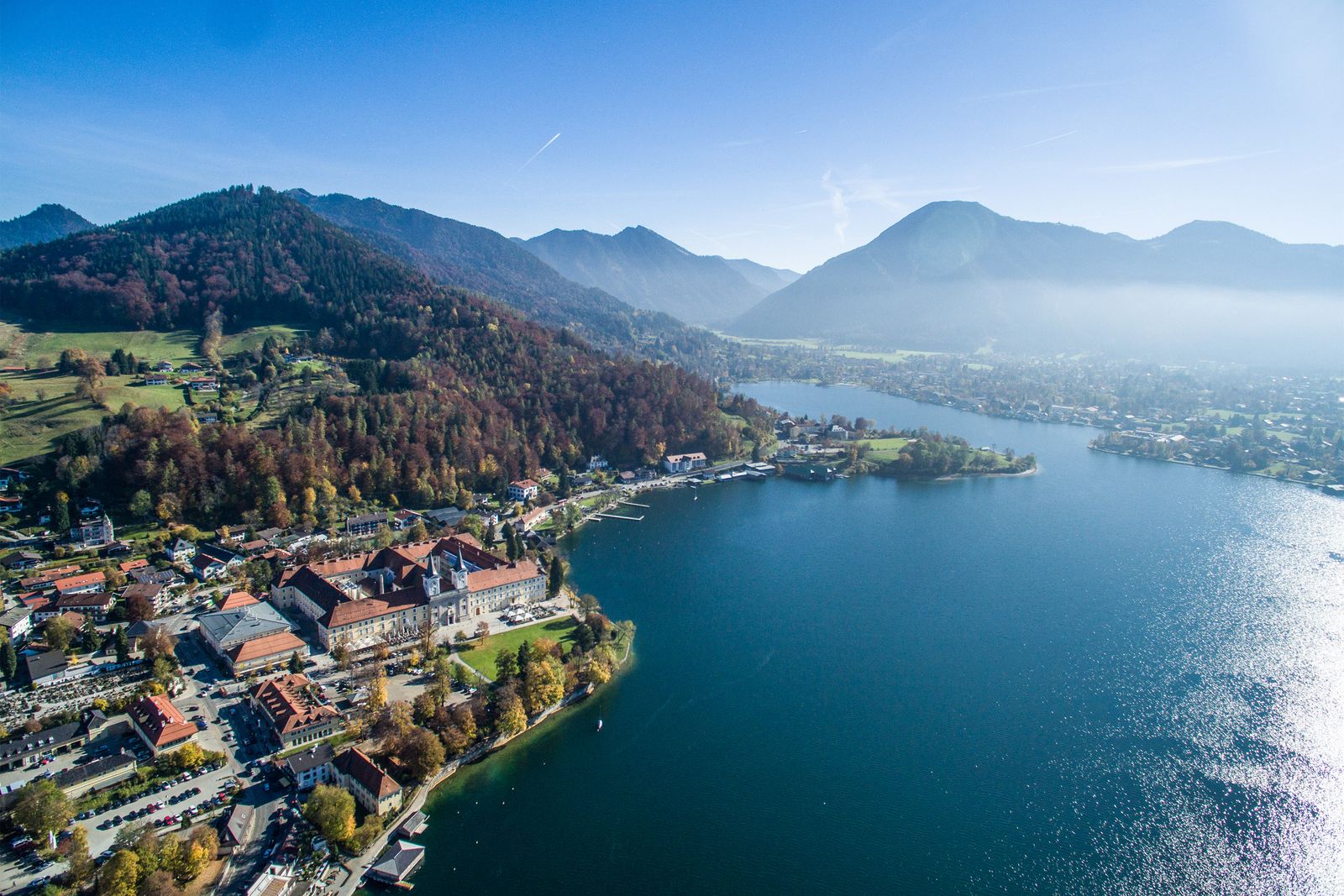 Aerial view over the Tegernsee with a village on the left shore, large pale buildings and autumn-coloured woods on the slope, deep blue water on the right and mountains in the haze behind