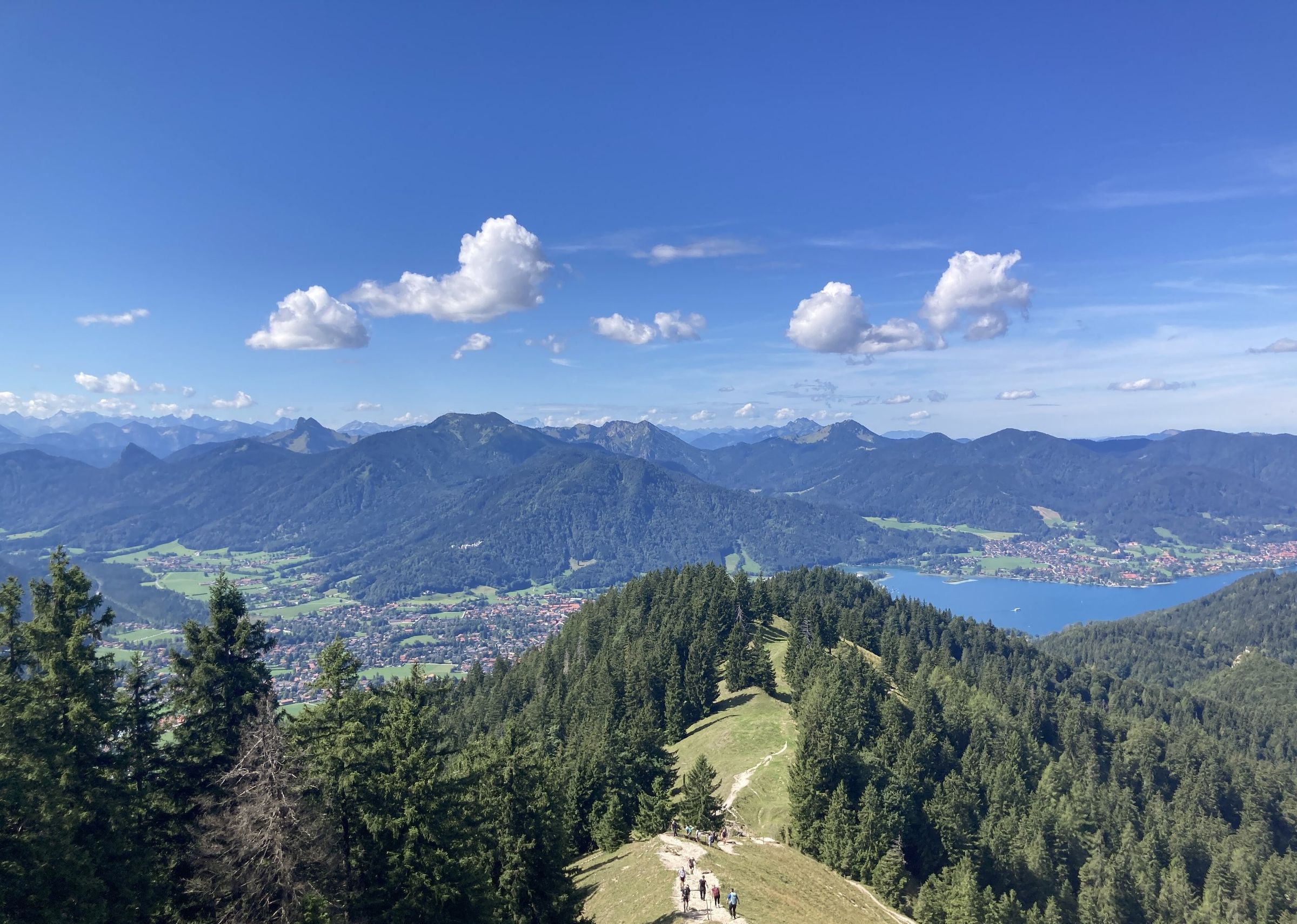 Wanderer klein auf einem Wiesengrat zwischen Bergwald, tief unten der Tegernsee mit Rottach-Egern, dahinter eine Alpenkette unter Quellwolken