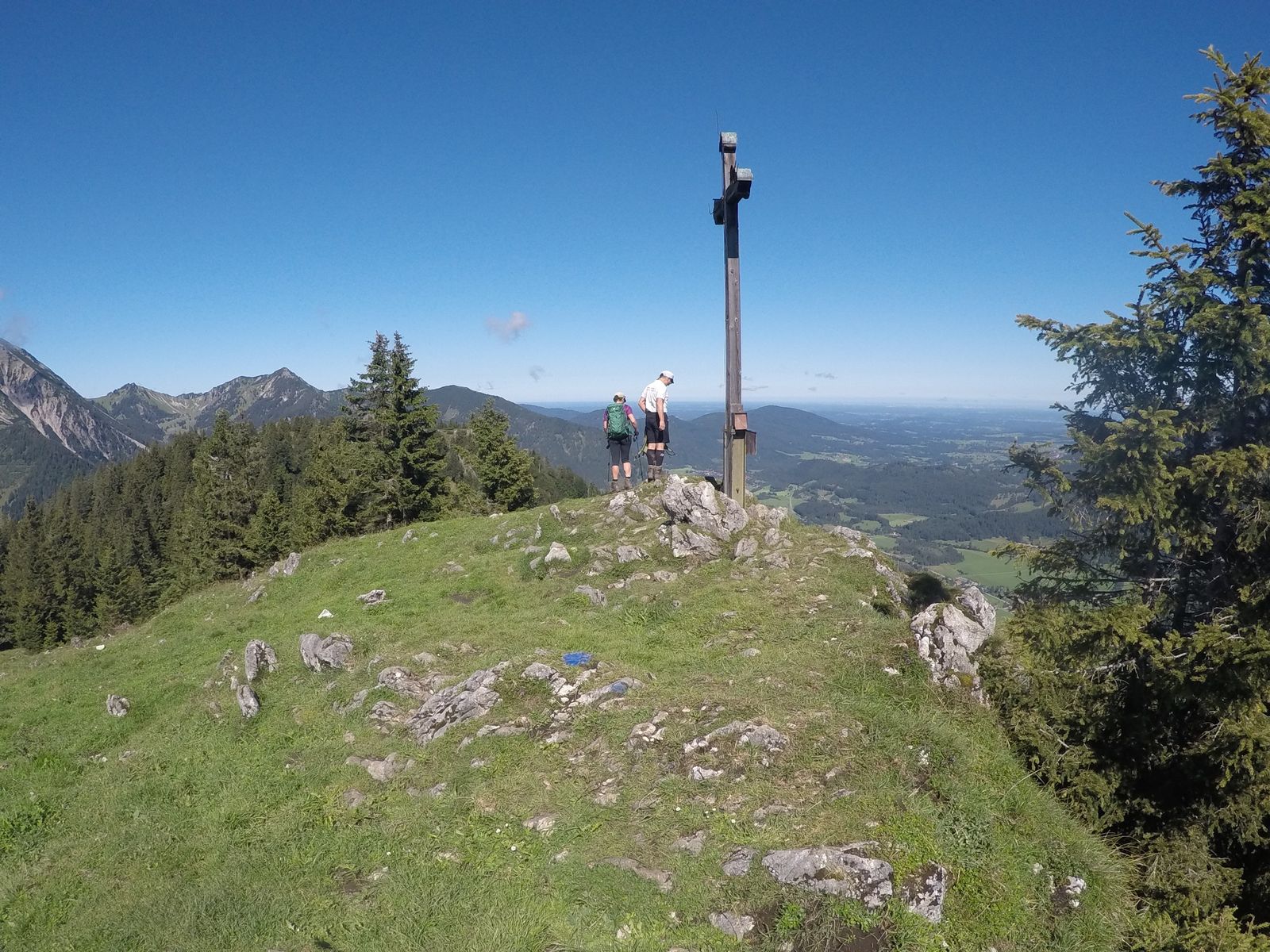 Zwei Wanderer von hinten neben einem schlanken Gipfelkreuz auf einer Graskuppe, Fichten am Rand, Blick ins Vorland und auf Berge