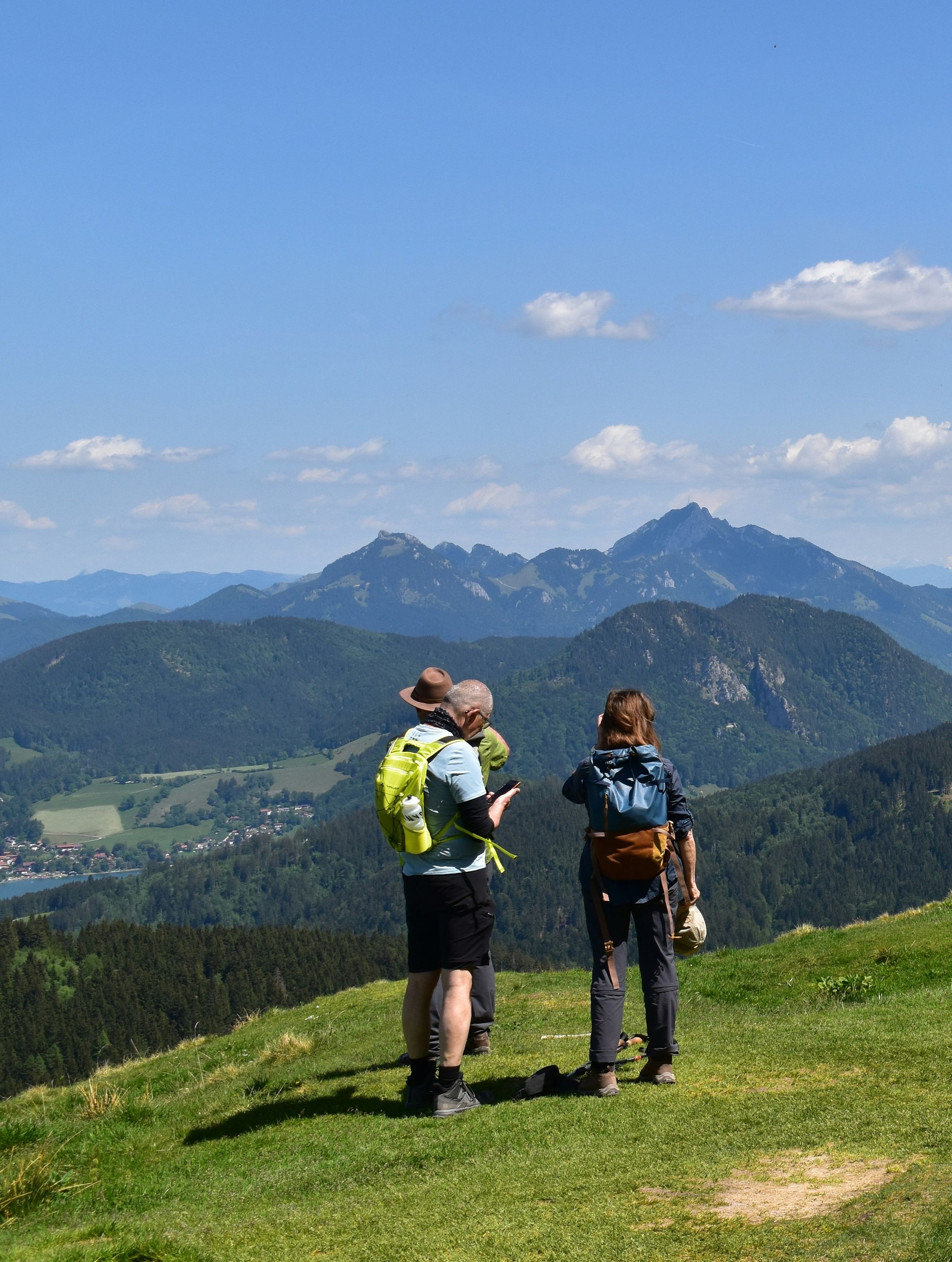 Drei Wanderer, zwei davon mit Rucksack, stehen von hinten gesehen auf einer Bergwiese und schauen über bewaldete Bergrücken und einen See mit Ort zu den Gipfeln am Horizont