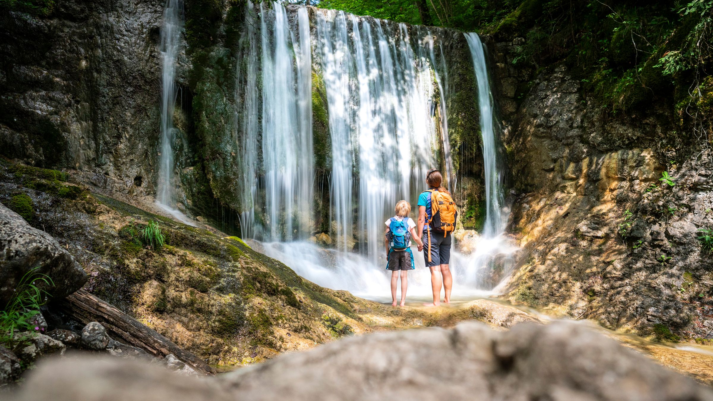 Ein Erwachsener und ein Kind mit Rucksäcken stehen von hinten vor einem breiten Wasserfall in einer Felsschlucht
