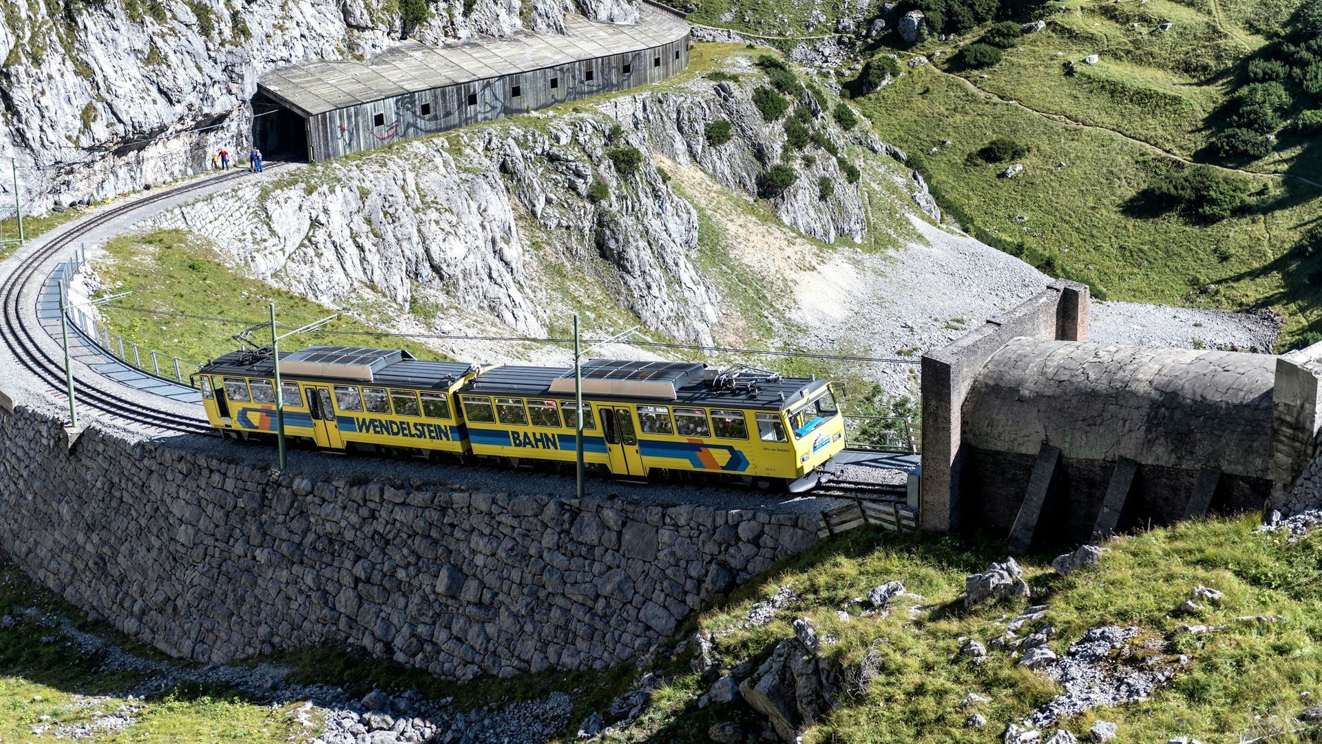 Gelber Triebwagen der Wendelsteinbahn in einer Kurve auf einer Stützmauer mit Galerie