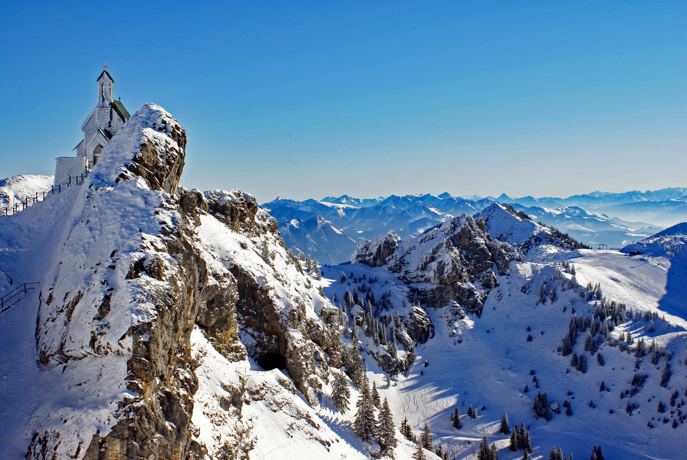 Die Kapelle auf einem verschneiten Felsen, dahinter eine weite Kette verschneiter Berge