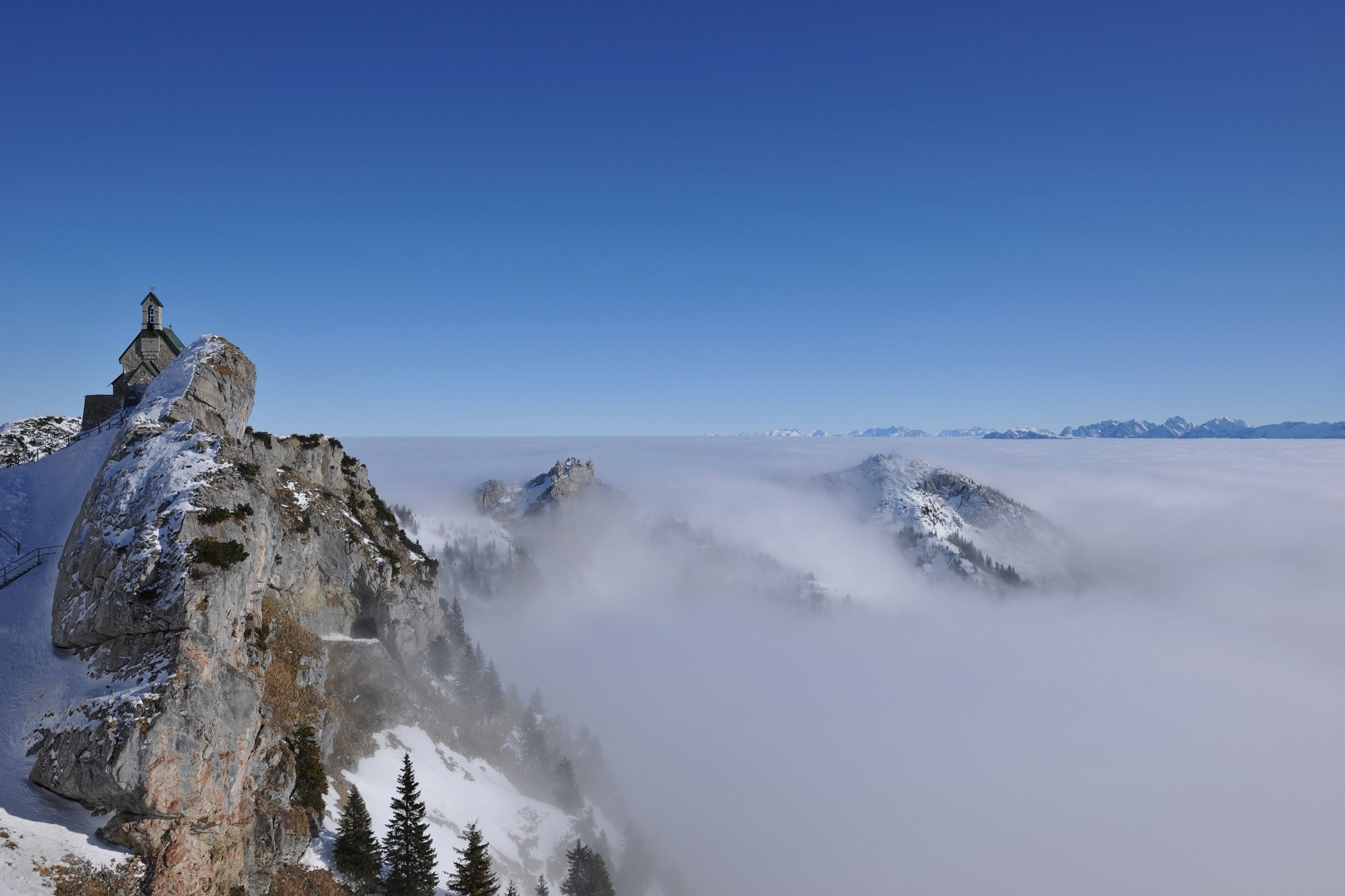 Die Kapelle auf dem Felsgrat ragt über eine geschlossene Nebeldecke, aus der einzelne Gipfel schauen