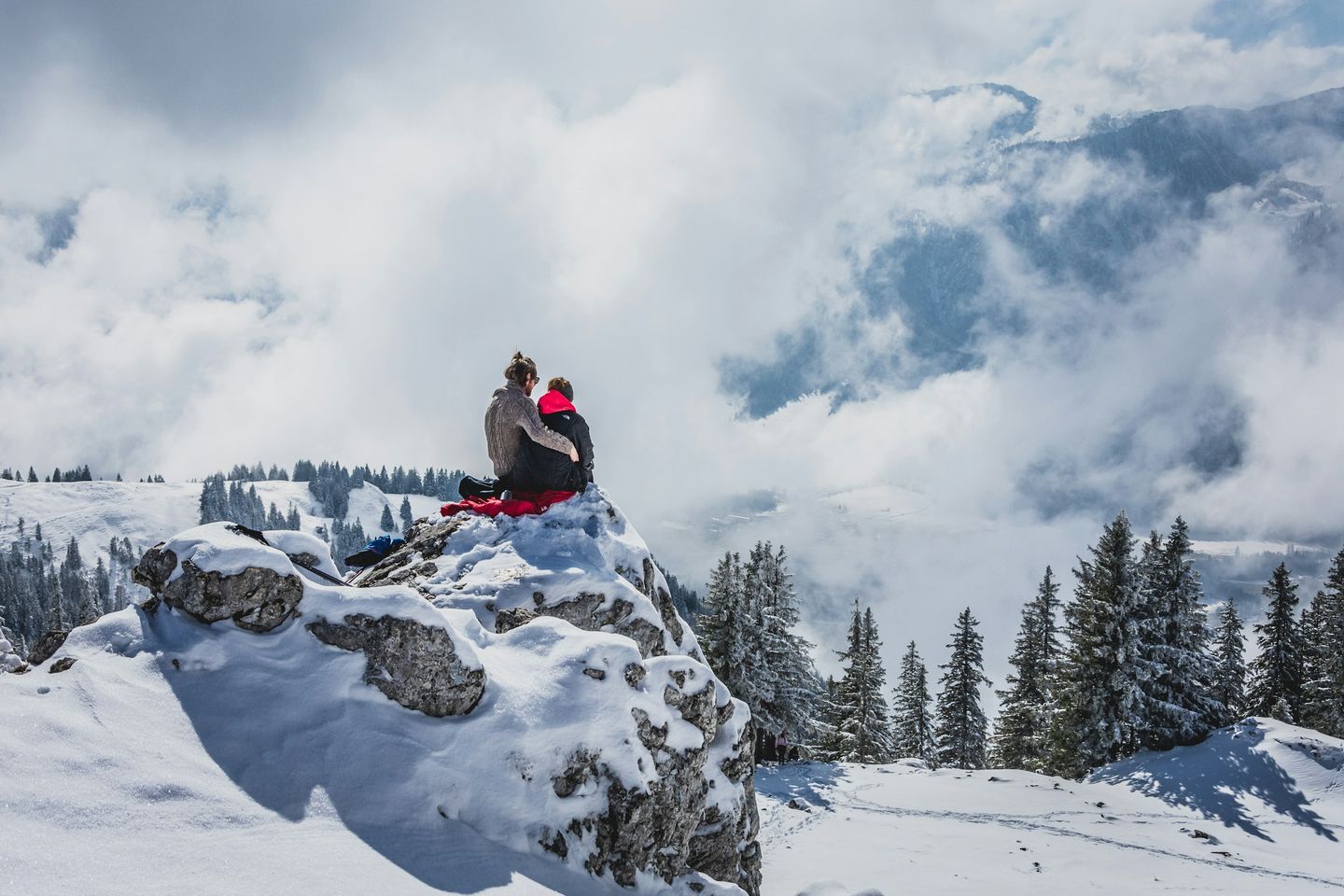 Zwei Personen sitzen von hinten auf einem verschneiten Felskopf, ringsum verschneite Fichten, Wolken und Nebel im Tal