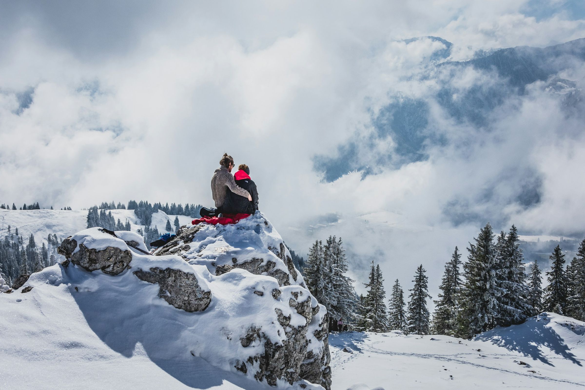 Zwei Personen sitzen von hinten auf einem verschneiten Felskopf, ringsum verschneite Fichten, Wolken und Nebel im Tal