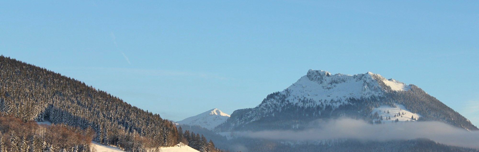 Verschneiter Felsgipfel über einem Nebelband, links ein verschneiter Waldhang, darüber blauer Himmel